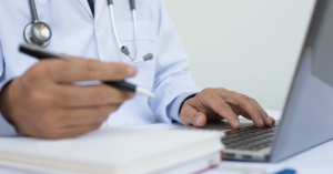 An unidentified general practitioner fills out paperwork in front of his laptop computer