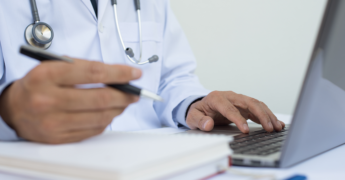 An unidentified general practitioner fills out paperwork in front of his laptop computer