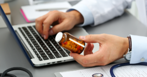 Male GP holds small jar of pills and types on laptop computer keyboard.